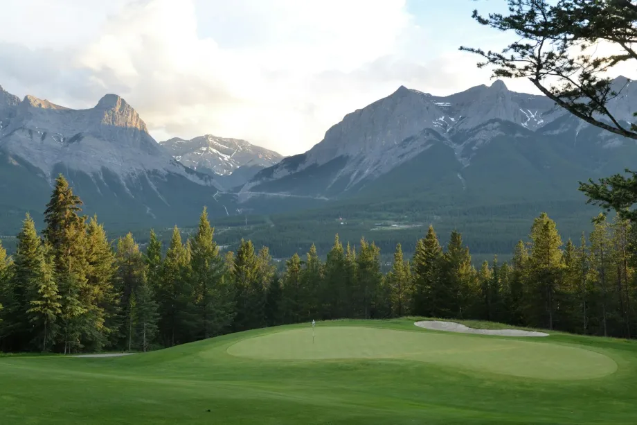 Mown golf green with pine trees below a rocky mountain and white clouds